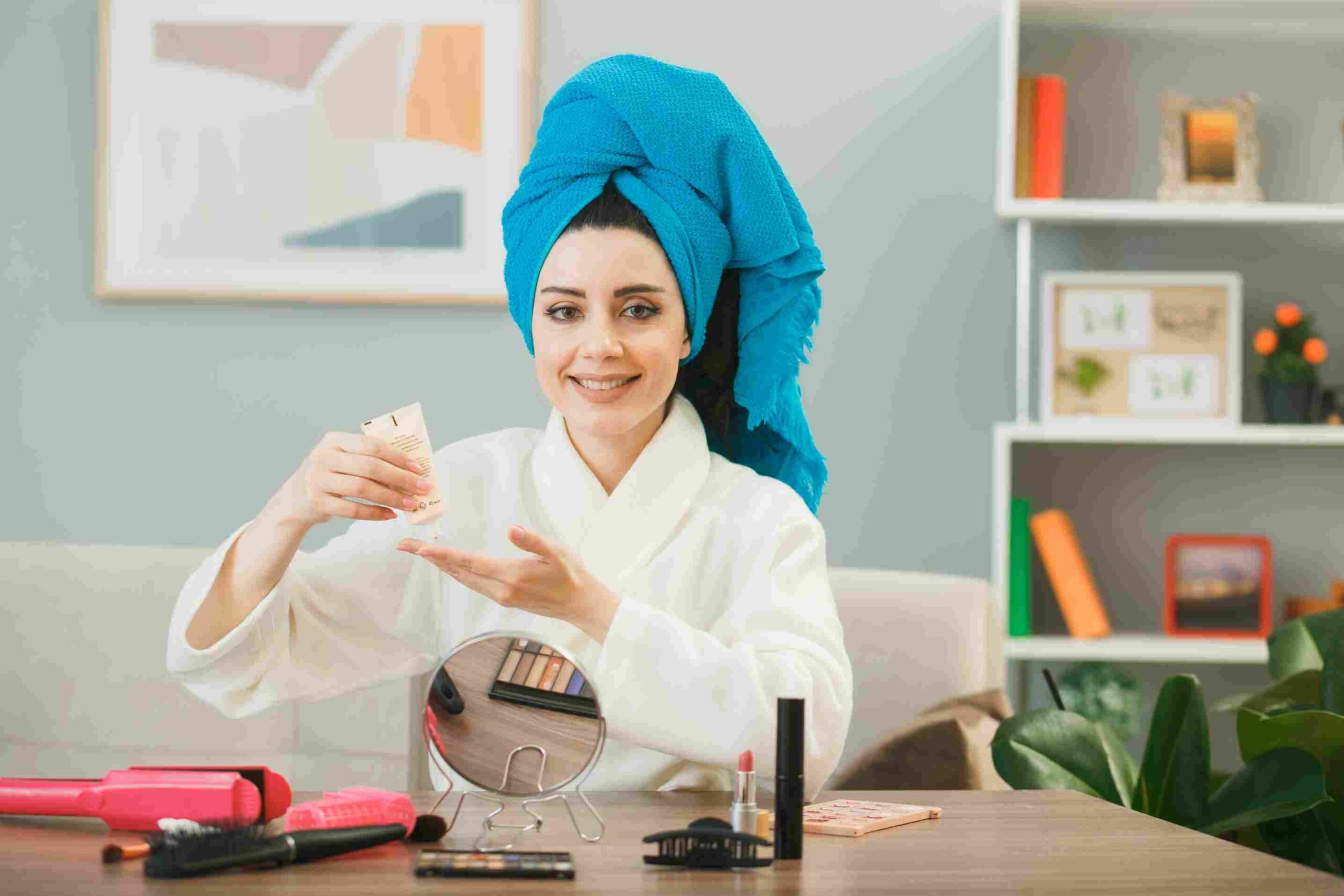 Smiling young girl holding tone-up cream wrapped hair in towel sitting at table with makeup tools in living room